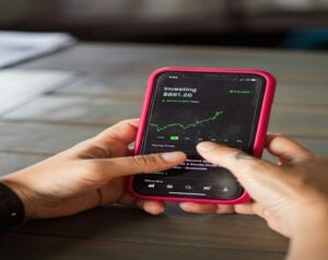 Hands hold a pink-cased smartphone displaying a stock trading app with a rising green chart and balance around $891.26 on a wooden table surface.
