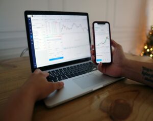 Person uses a laptop with a trading dashboard while holding a smartphone showing stock charts in a trading app
