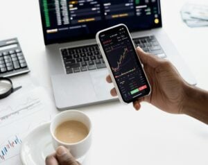 Person holds a smartphone showing a stock chart and market data, with a laptop and charts on the desk behind.