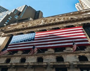 A large American flag draped across the stone front of a grand classical building, with smaller flags and modern skyscrapers in the background.